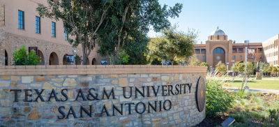 Photograph of Texas A&M San Antonio courtyard with grass and several stone buildings and the university name and logo displayed on a large stone dias at the forefront of the photograph.