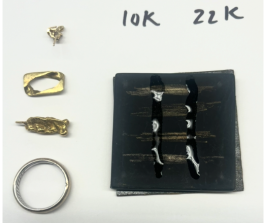 A photograph of four pieces of gold jewelry and a black slate which they have been drawn across. On top of the metal scratches are acids showing that some of the items are not gold, and some are atleast 10k gold.  
