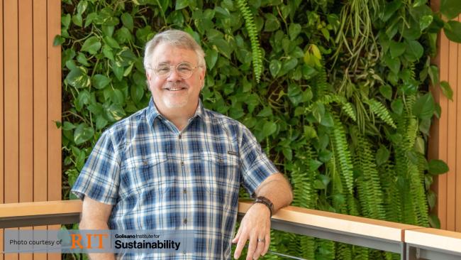 A white man in a blue plaid shirt smiles, standing in front of a wall covered in vibrant green ferns. Small white text reads: ask nature, hive-- the text is punctuated by a small hexagonal pattern.